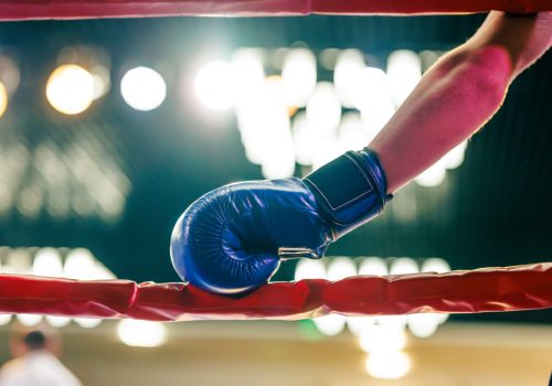 A close-up of a boxer holding a red boxing glove, wearing blue shorts, in a boxing ring. The background features bright lights, creating an intense atmosphere.