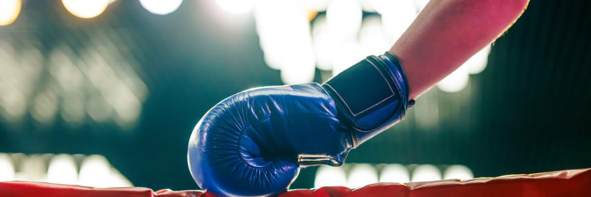 A close-up of a boxer holding a red boxing glove, wearing blue shorts, in a boxing ring. The background features bright lights, creating an intense atmosphere.