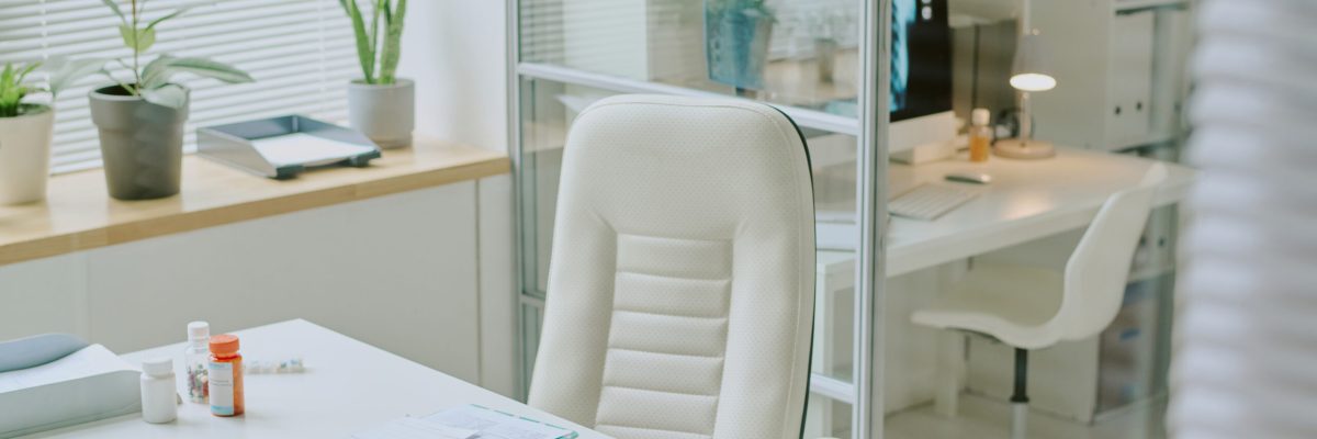 Bright and airy medical office with white furniture, green plants, and various medical equipment arranged on desk and in the room. Shelves and glass partitions adding to organized look